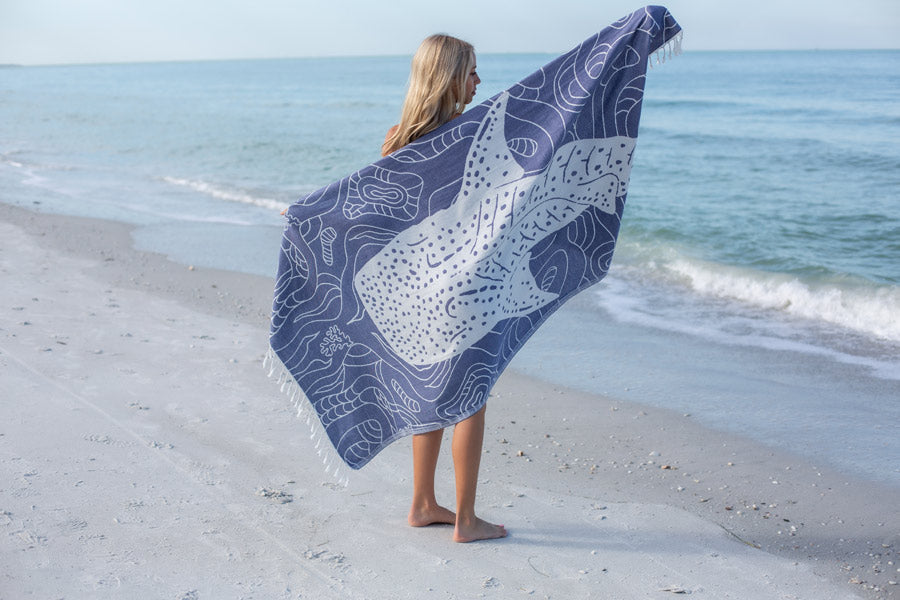 Person standing on a beach with a blue and white patterned towel draped over their shoulders.