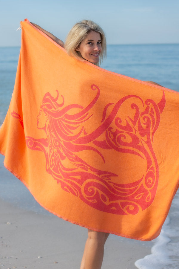 Woman holding an orange towel with a red dragon design on a beach.