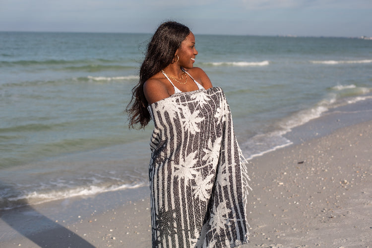 Woman standing on a beach with a gray and white floral patterned towel draped over her shoulders.