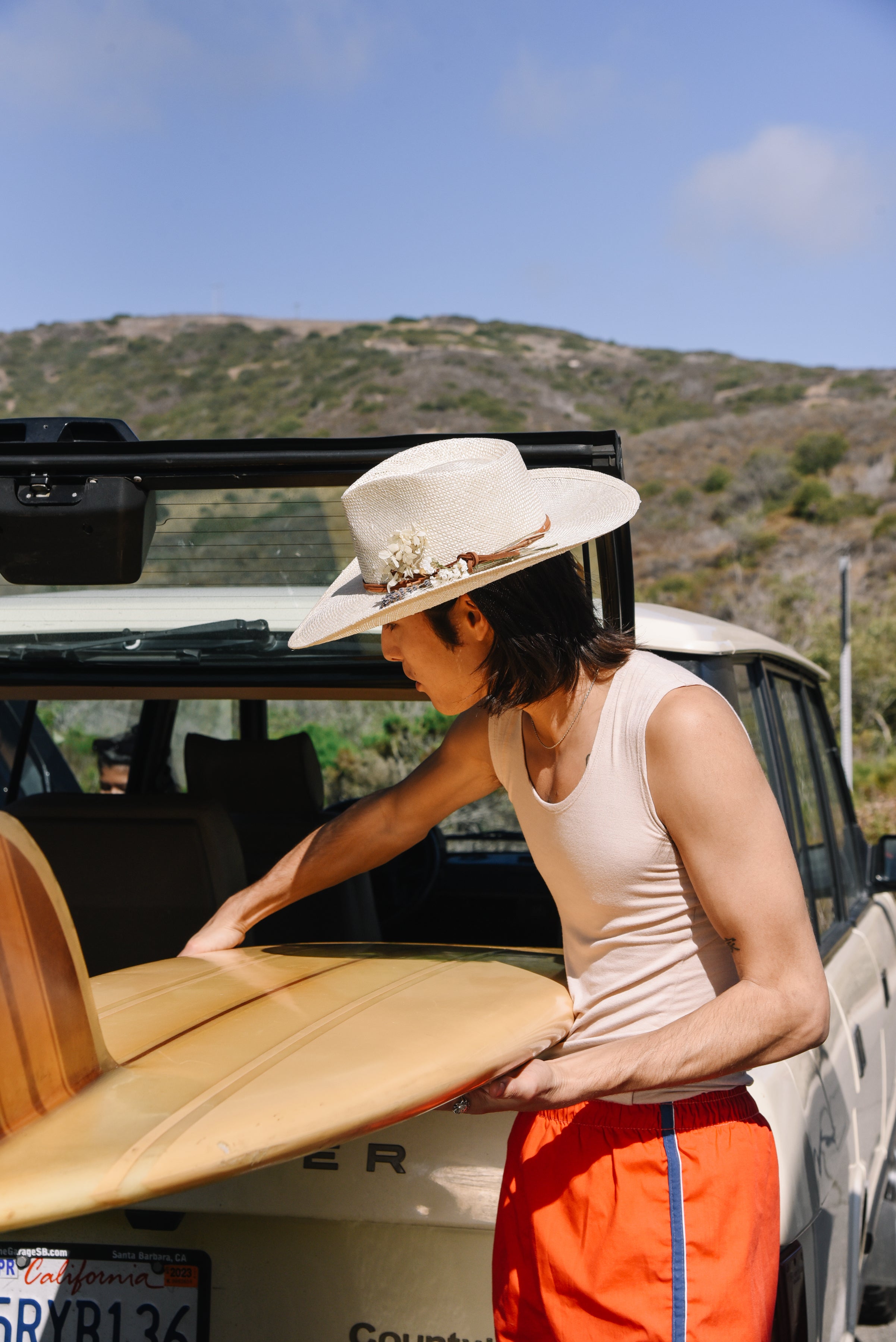 Person with a surfboard standing next to an open car trunk in a scenic area