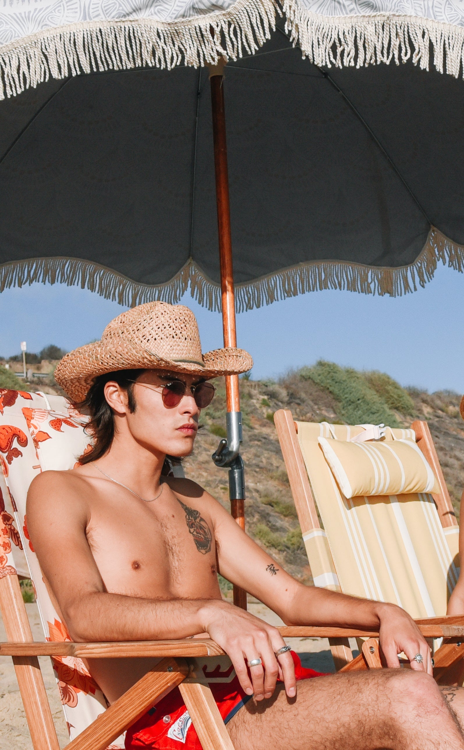 Man sitting under a large black umbrella on a beach chair, wearing a straw hat and sunglasses.