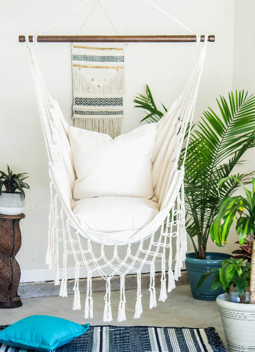 White macrame hammock chair with cushions in a room with plants and a rug.