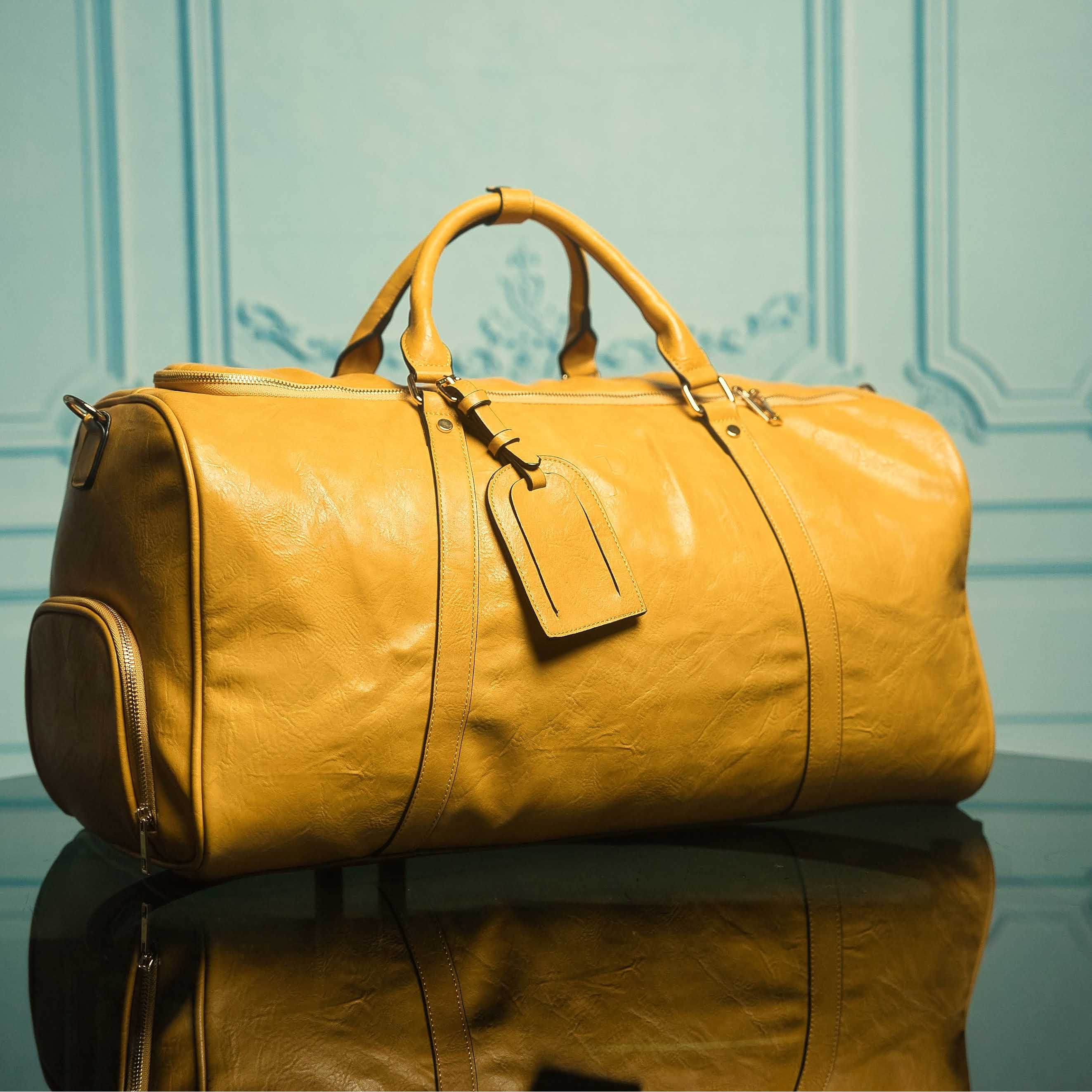 Yellow leather duffel bag with a tag on a reflective surface and light blue background