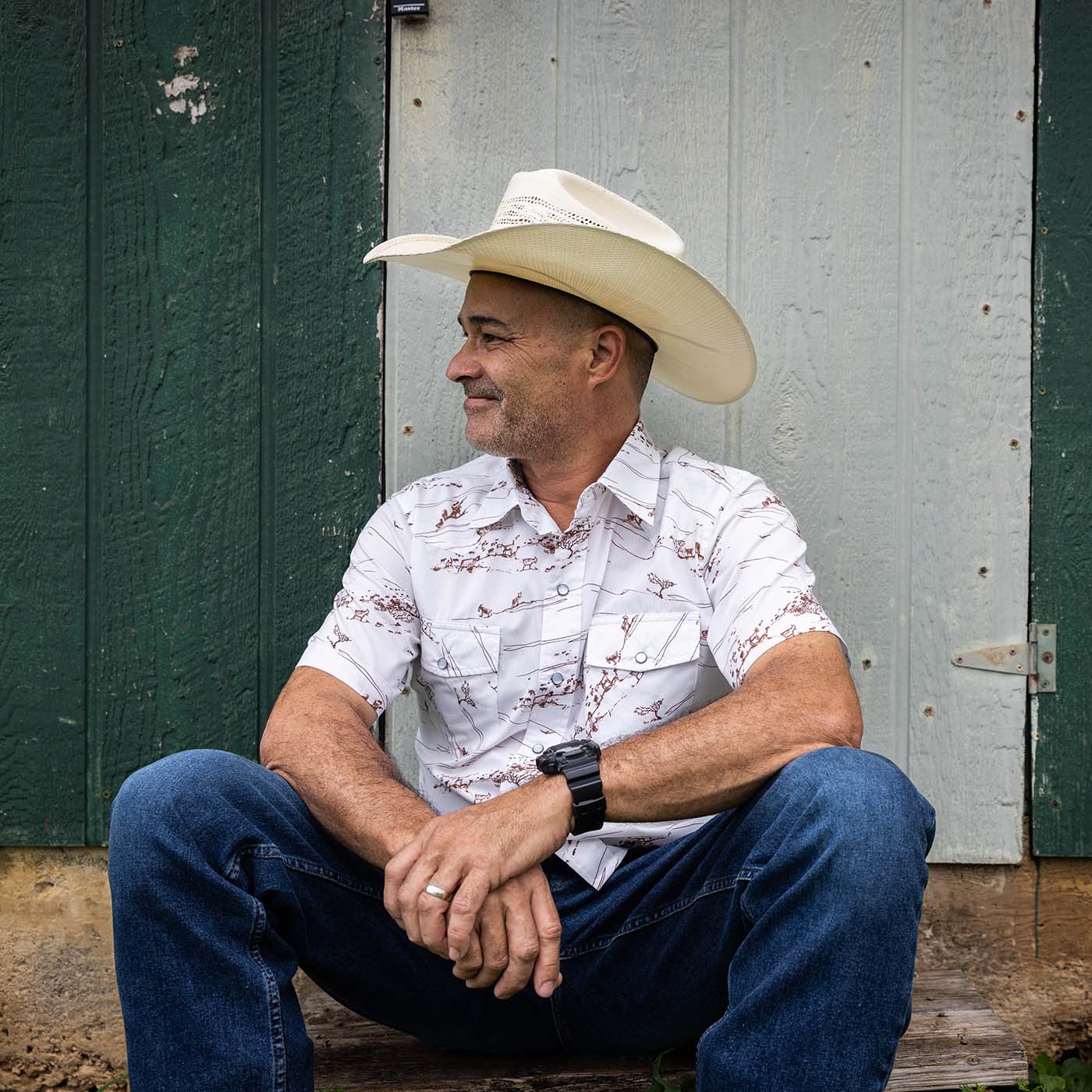 Man wearing a cowboy hat and white shirt sitting against a green wooden wall.