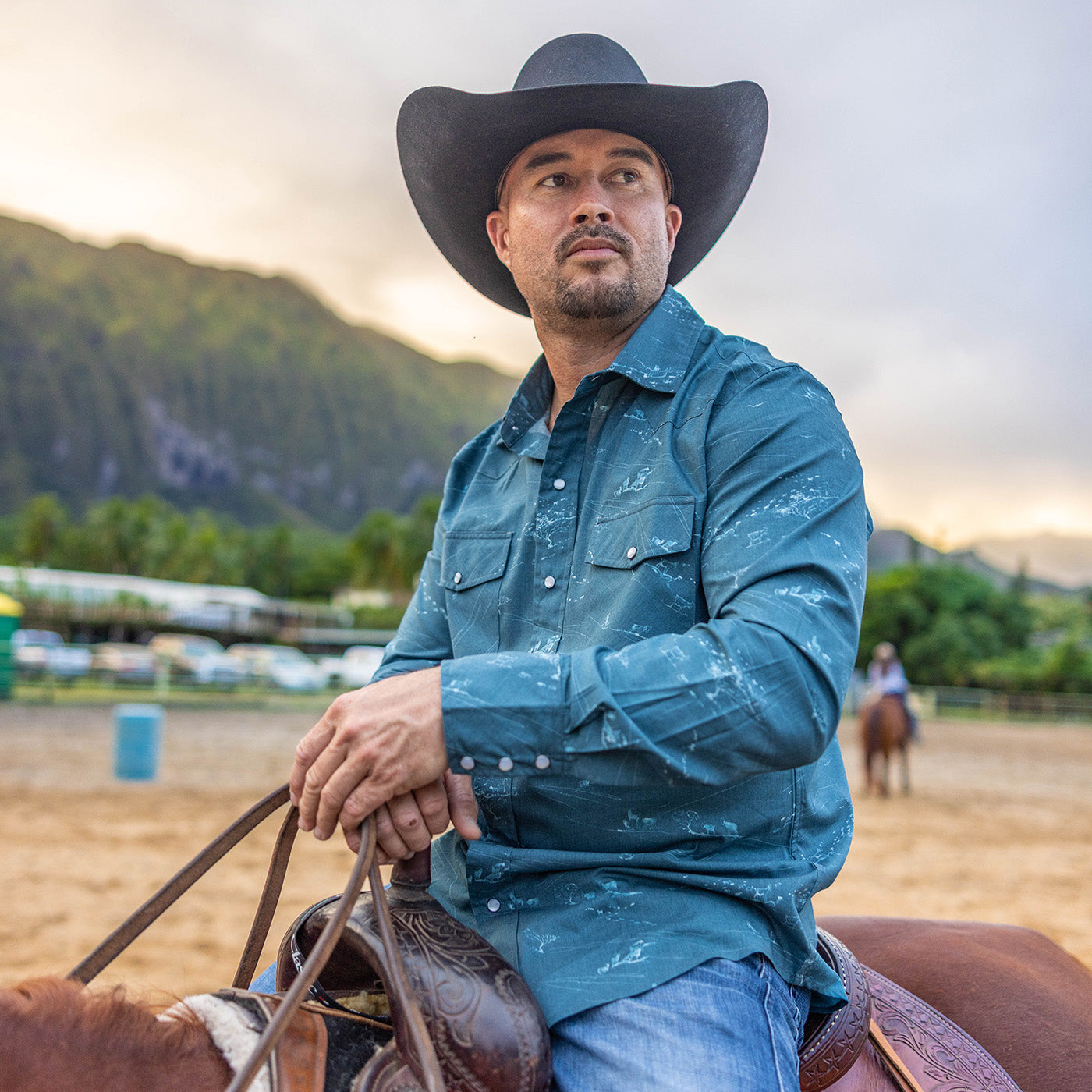 Man in a cowboy hat and blue shirt sitting on a horse with mountains in the background