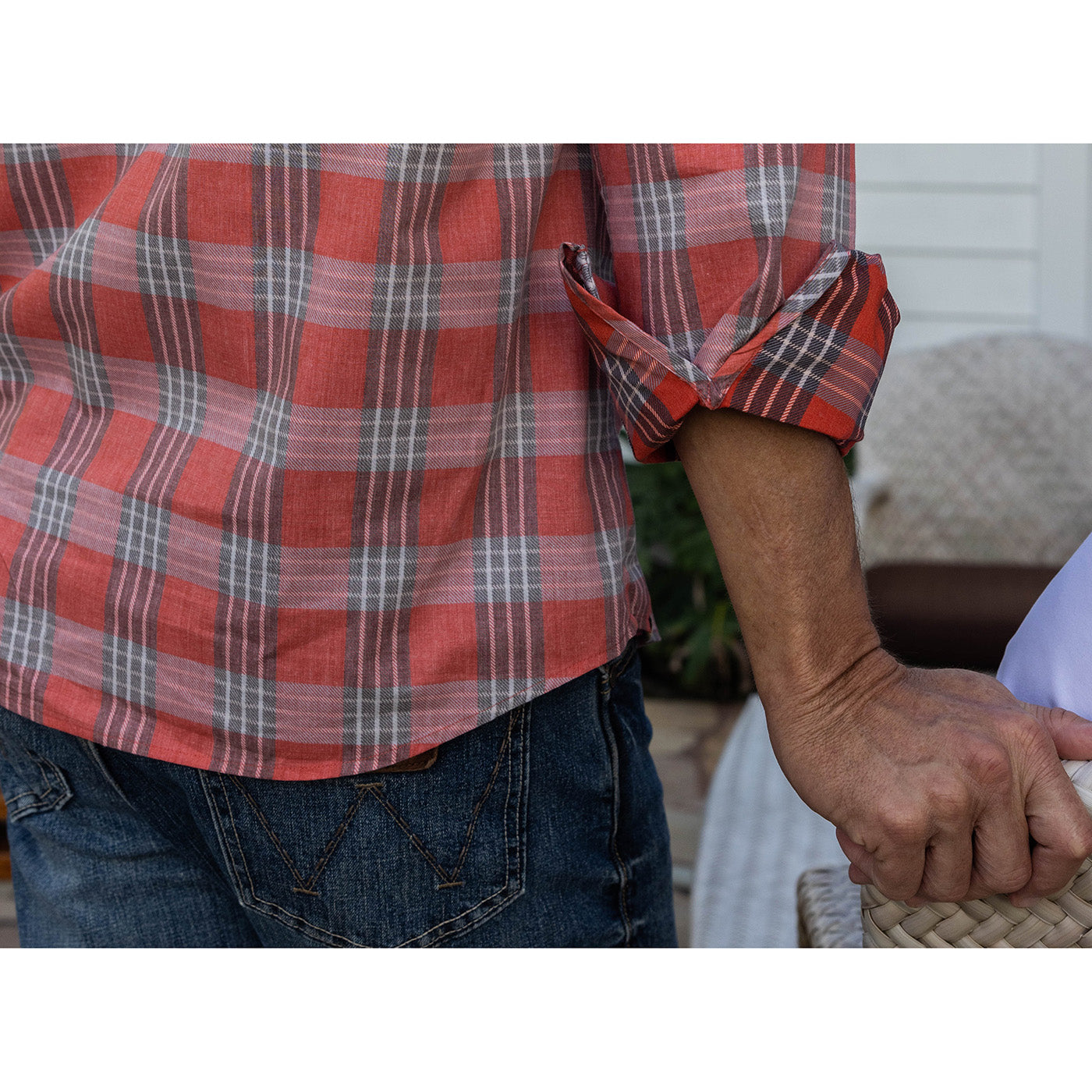 Person wearing a red plaid shirt and blue jeans, holding a woven bag.