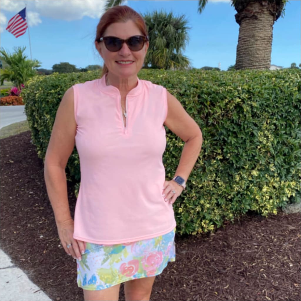 Woman wearing a pink sleeveless top and floral skirt standing outdoors with greenery and a palm tree in the background.
