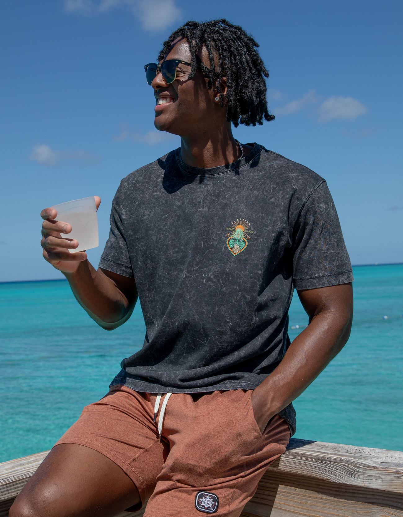 Man sitting by the ocean wearing a dark t-shirt with a logo and sunglasses.