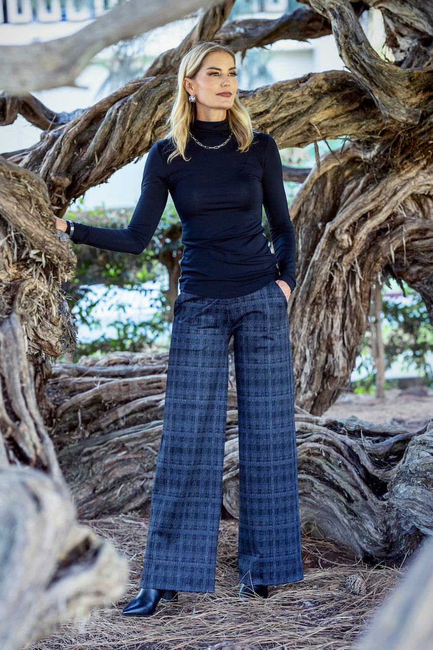 Woman in a black top and blue plaid pants standing among large tree roots.