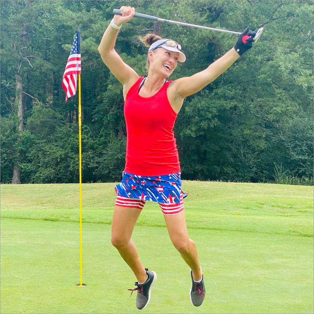 Woman in red tank top and patriotic shorts celebrating on a golf course with an American flag in the background.