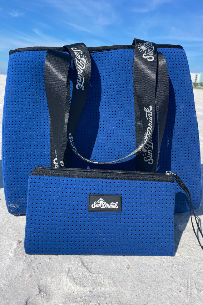 Blue textured tote bag with black strap and visible brand logo on a sandy background