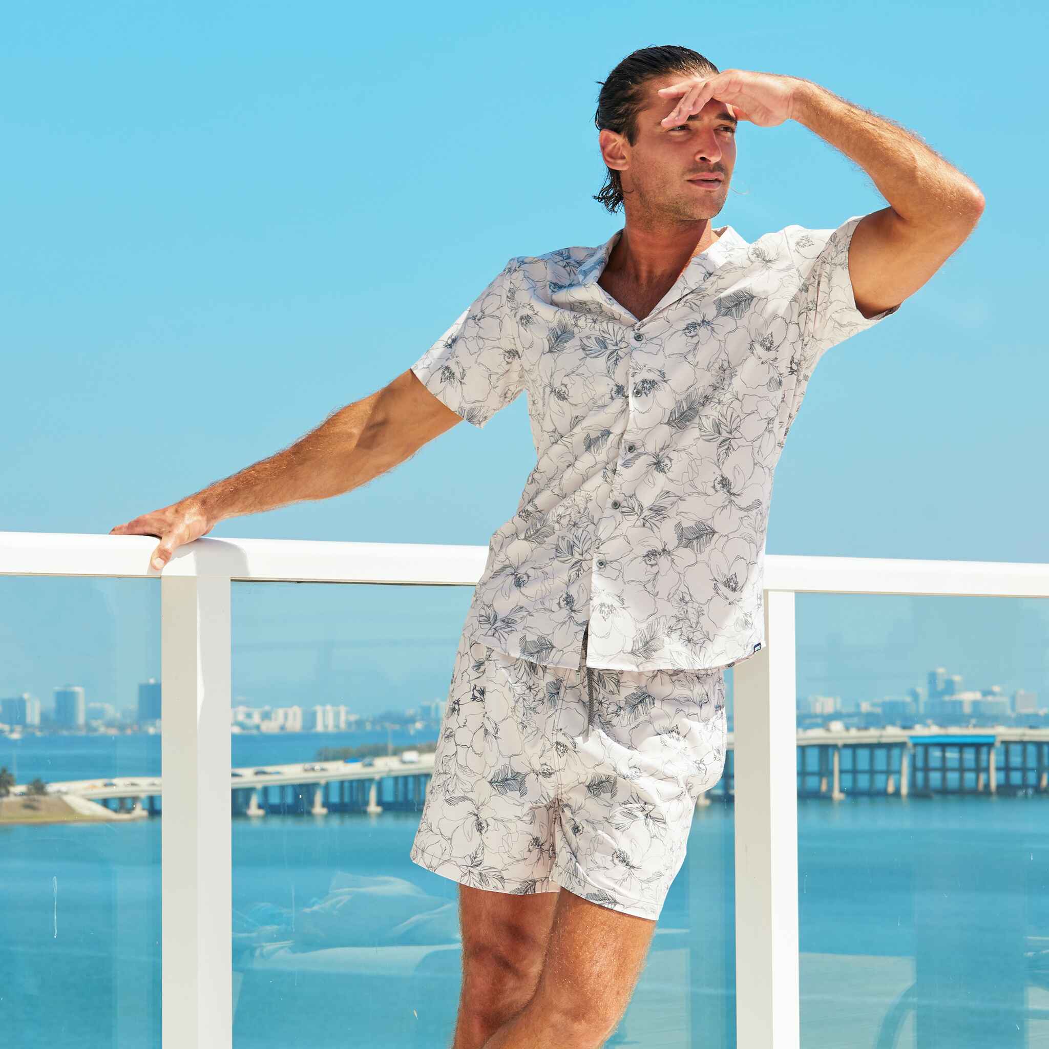 Man in a light-colored floral shirt and shorts standing on a balcony with a cityscape in the background.