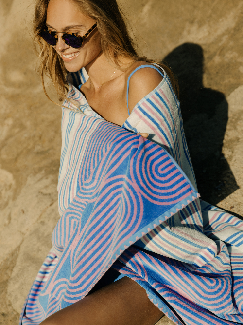 Woman wearing a colorful striped dress with a patterned towel draped over her shoulders against a beige wall.