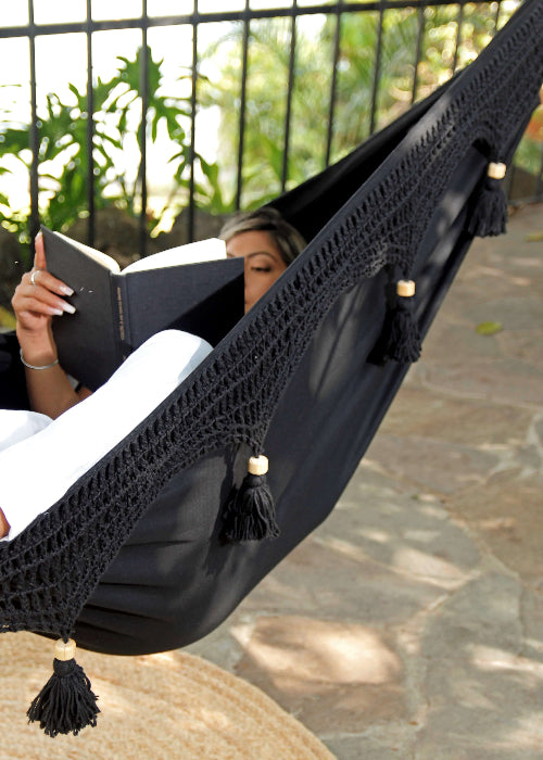 Person reading a book in a black hammock with tassels on a stone patio.