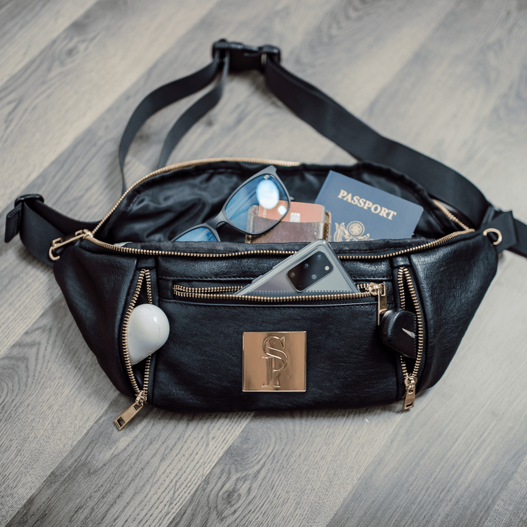 Black waist bag with gold accents on a wooden floor, containing items like a passport and phone.