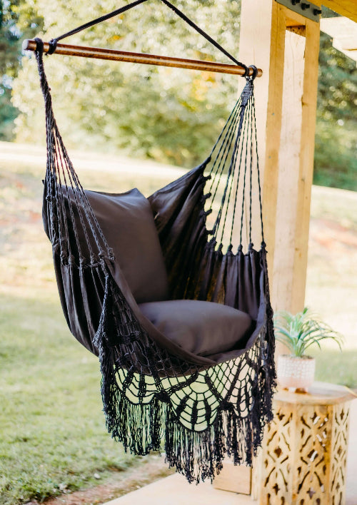 Black hammock chair with fringe hanging outdoors on a wooden stand.