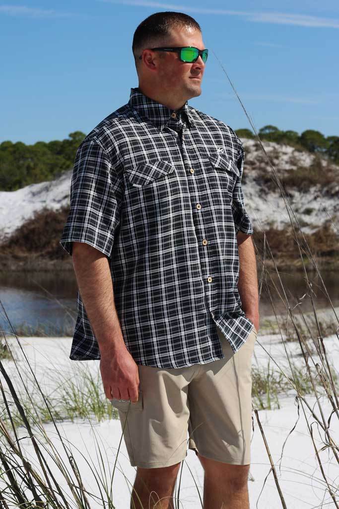 Man wearing a plaid shirt and sunglasses standing on a beach with a scenic background.