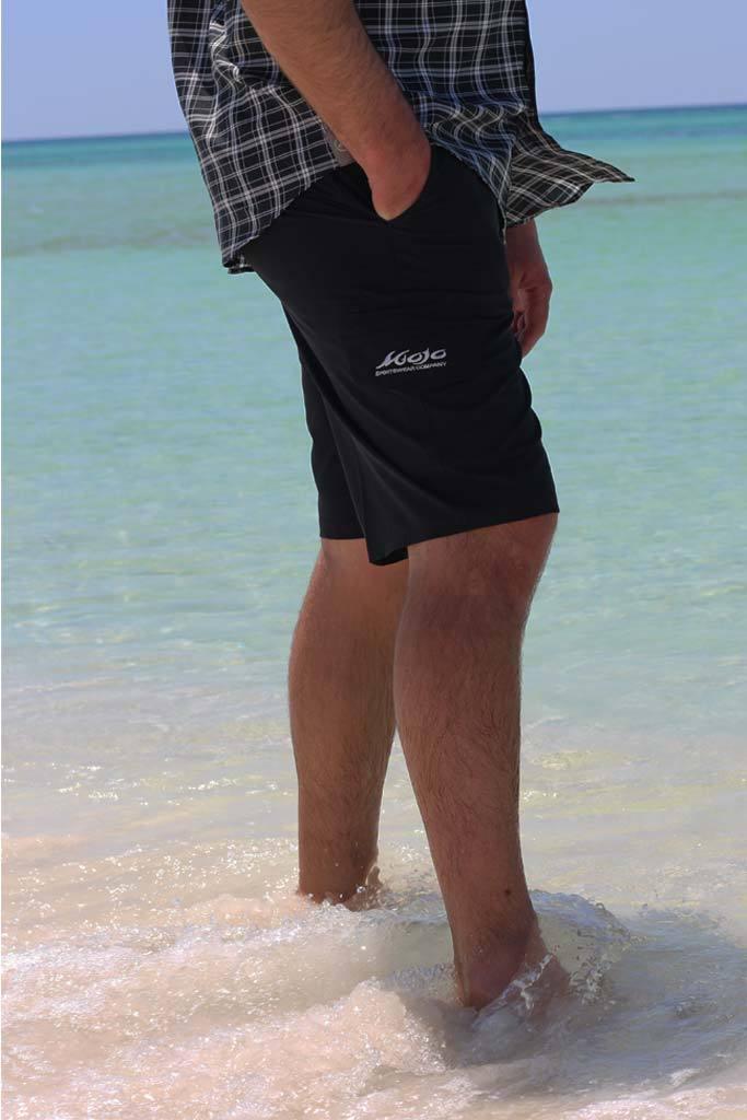 Person wearing black shorts with a logo standing in shallow water at the beach.