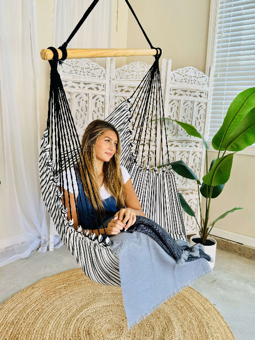 Woman sitting in a striped hammock chair in a home setting with a plant and rug.