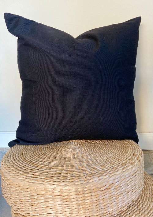 Black pillow on a woven stool against a white wall