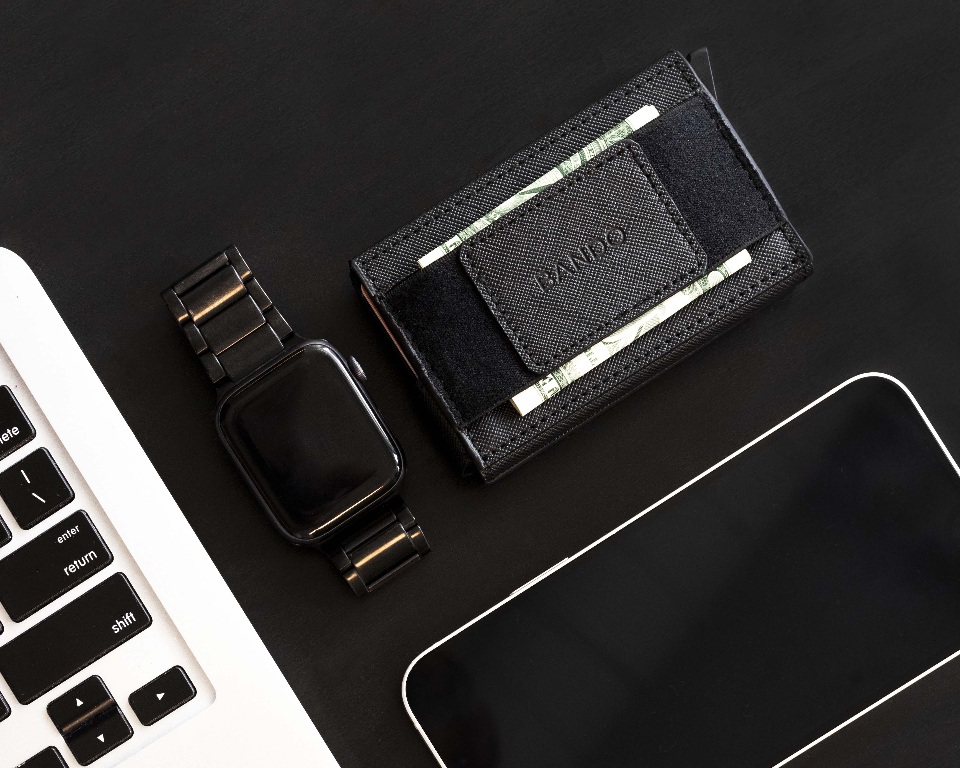 Black wallet with money clip and smartwatch on a black surface with computer keyboard and phone.