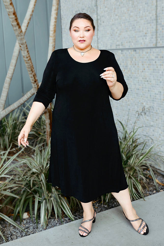 Woman wearing a black dress standing outdoors with plants and a wall in the background