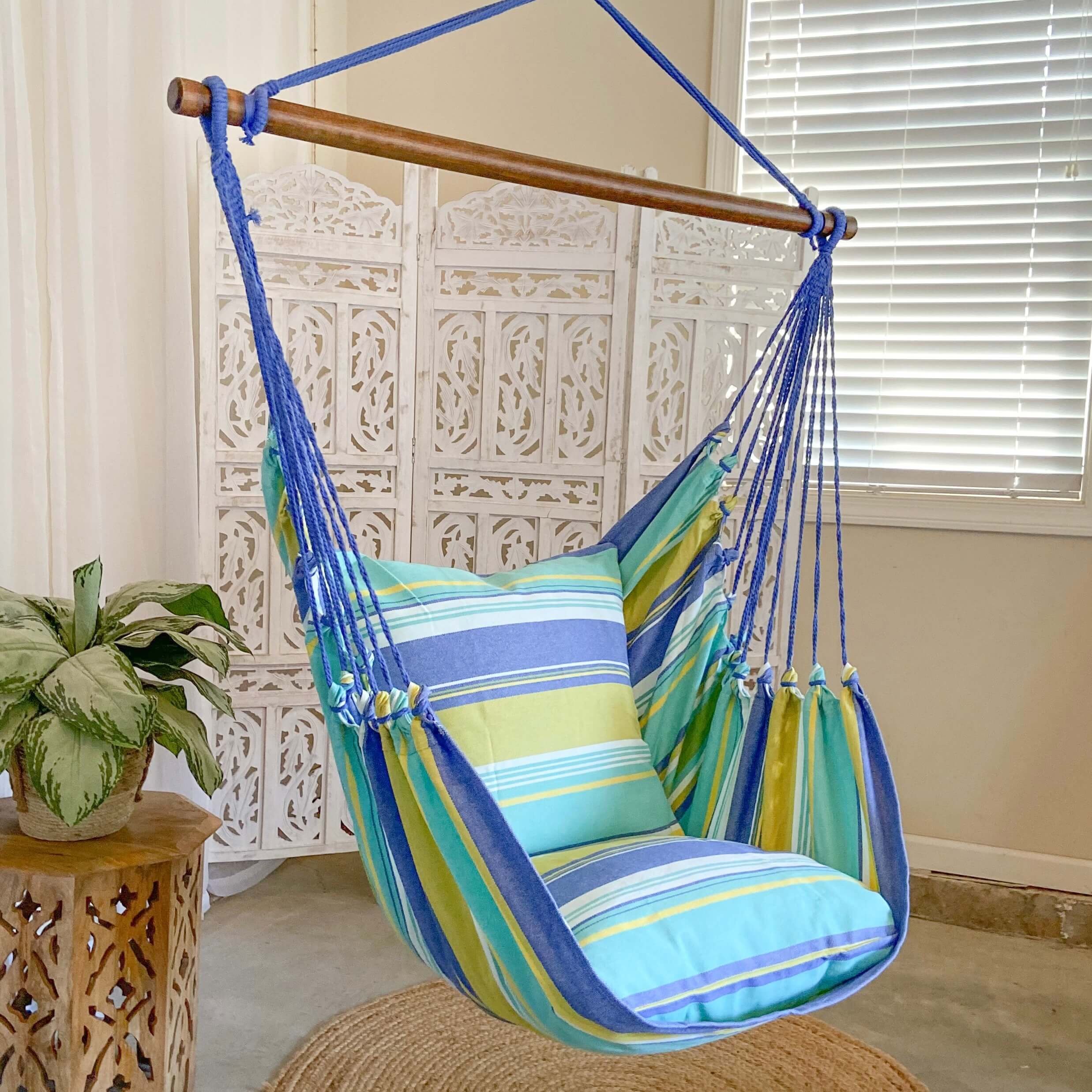 Colorful striped hammock chair hanging indoors with a plant and table in the background.