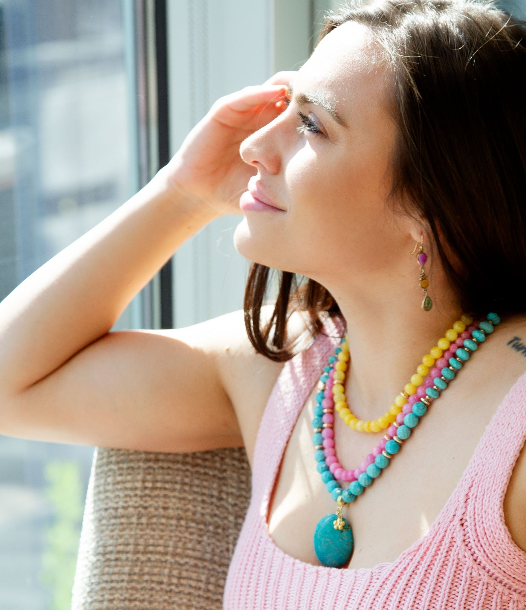 Woman wearing colorful beaded necklaces and earrings, standing by a window.