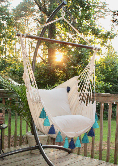 Beige hammock chair with blue tassels on a wooden deck during sunset.
