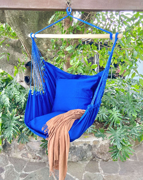 Blue hammock chair with a brown blanket hanging on a tree branch with green leaves in the background.