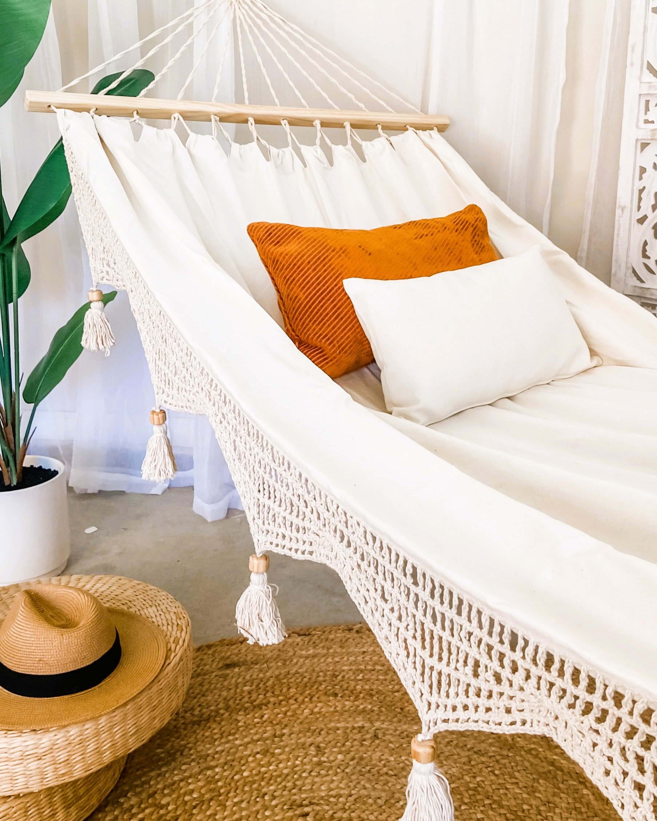 White macrame hammock with orange and white cushions in a room with a plant and woven chair.