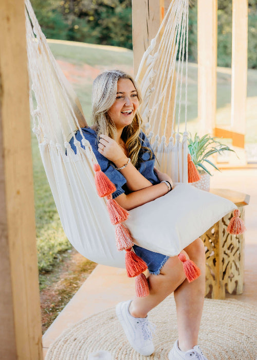 Woman sitting in a hammock with tassels, smiling outdoors.