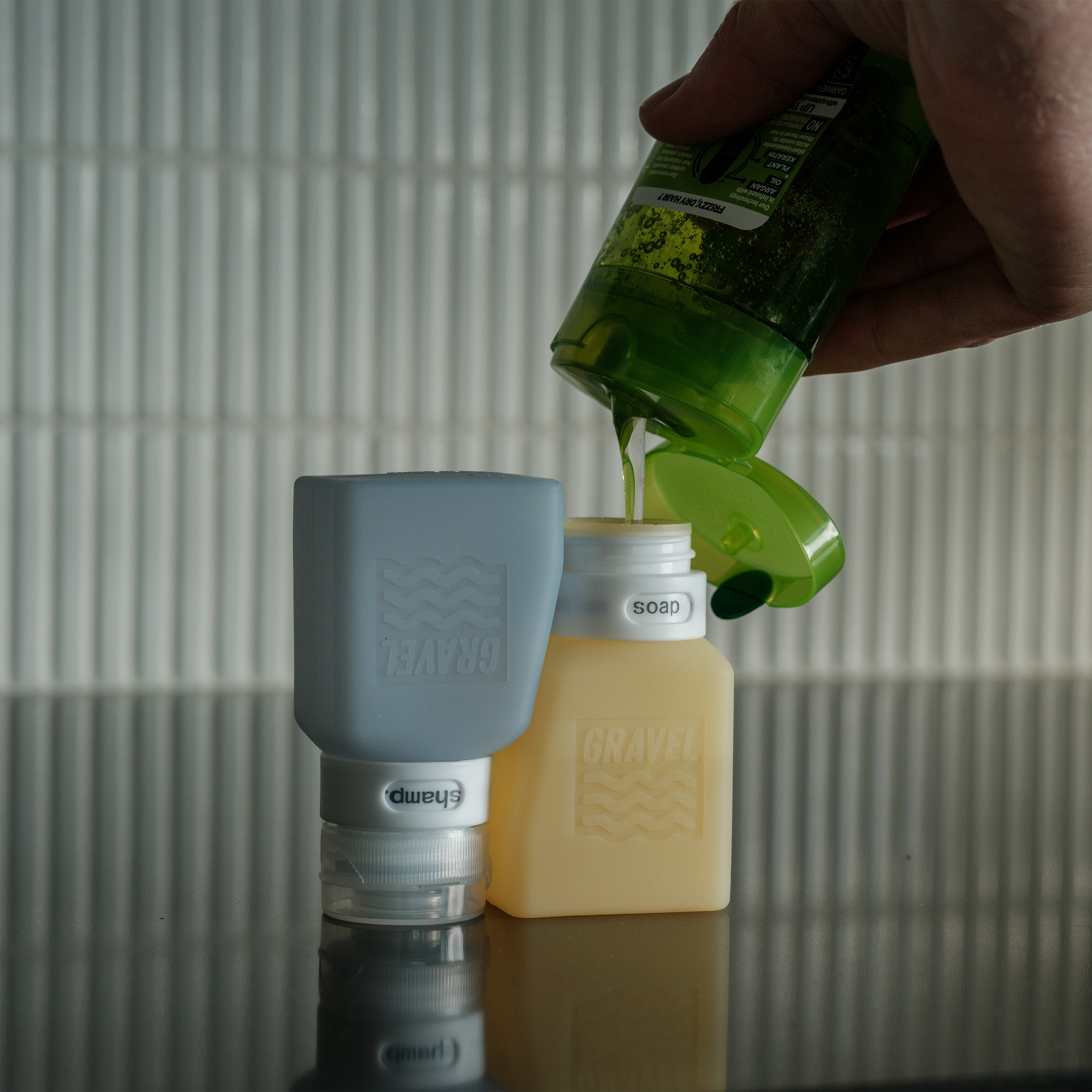 Person pouring a green liquid from a bottle into a blue container on a reflective surface.