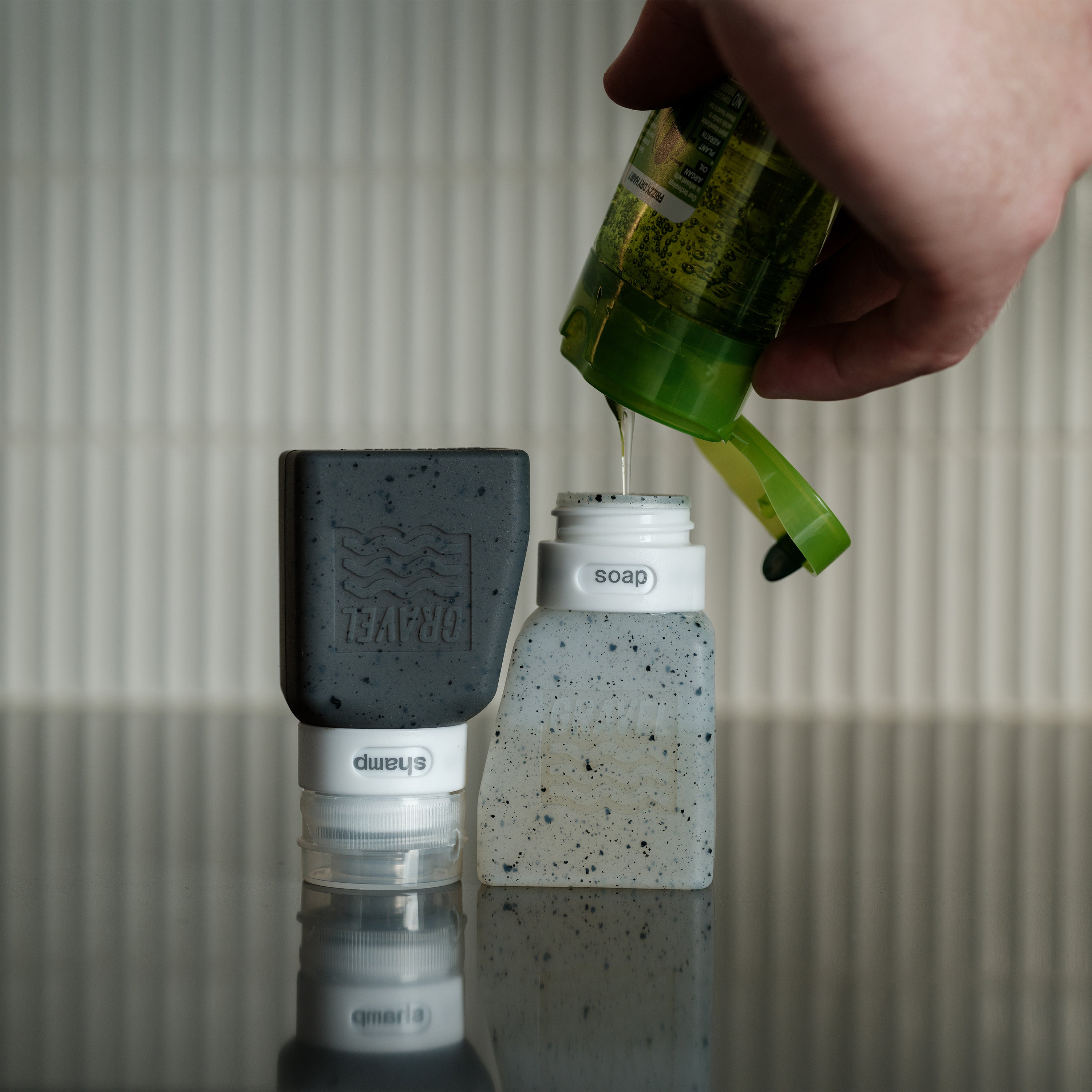 Hand pouring soap from a green bottle into a concrete soap dish with a black scrubber on a neutral background.