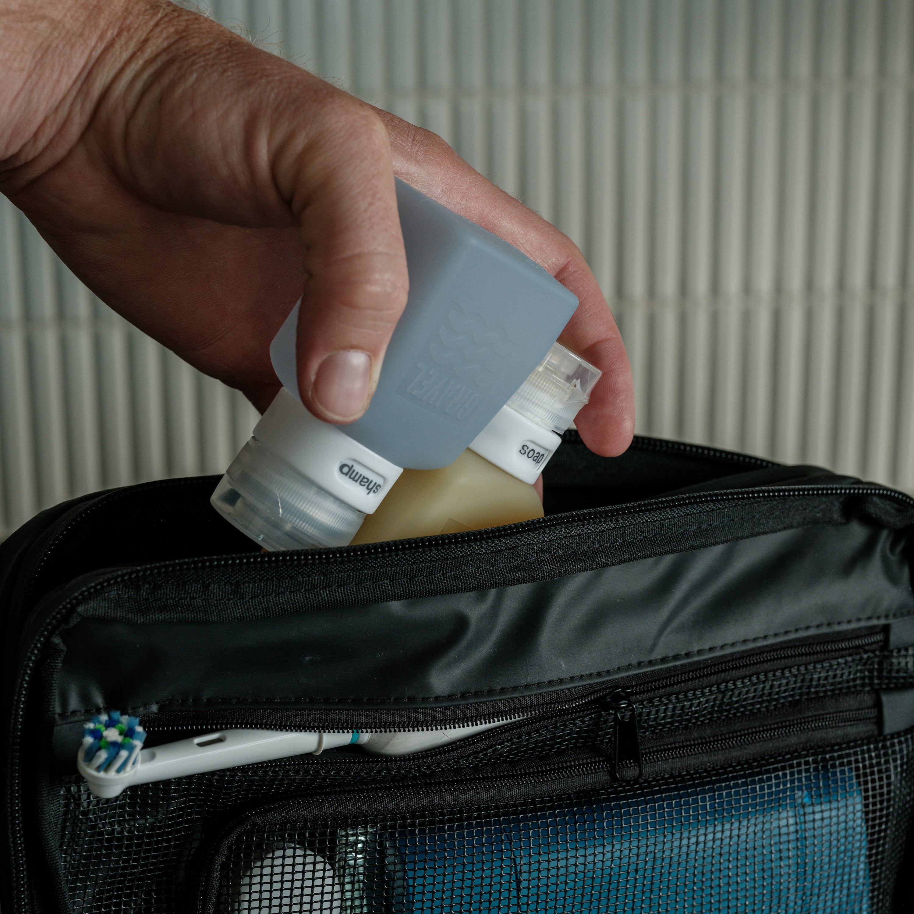 Hand placing a bottle into a black toiletry bag with a toothbrush visible.