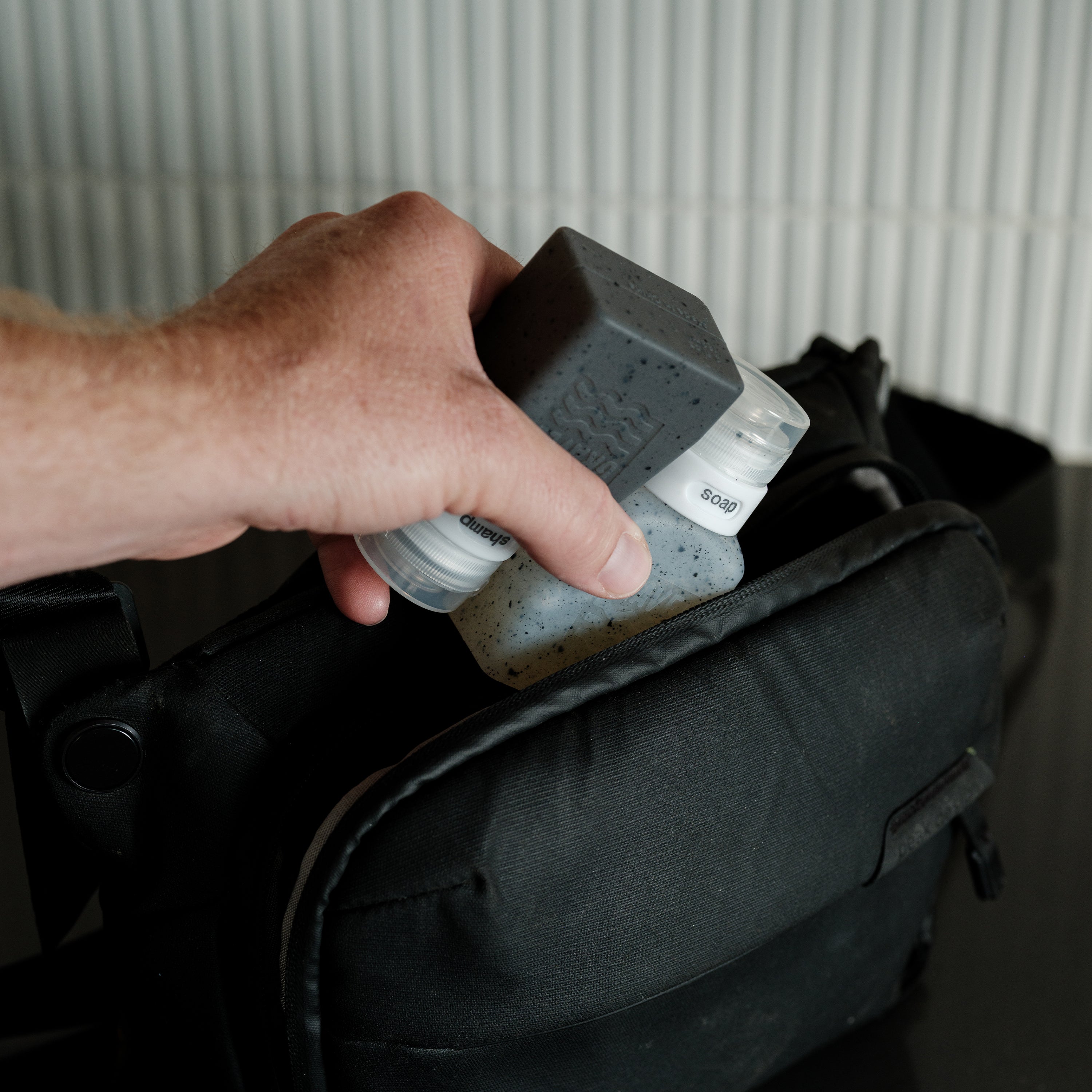 Hand holding a black and gray container over a black bag with a white background