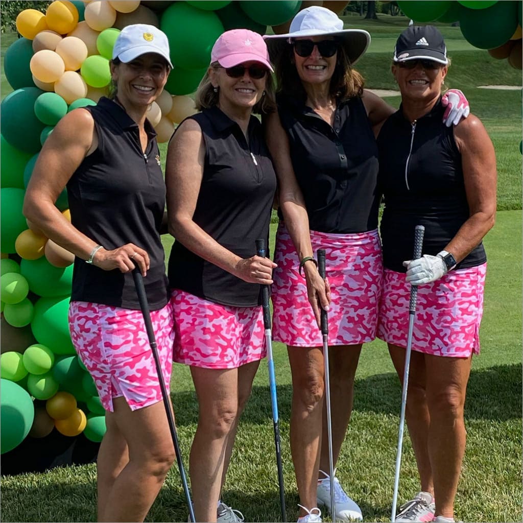 Four women in golf attire with pink shorts and black tops, standing in front of a colorful balloon arch.