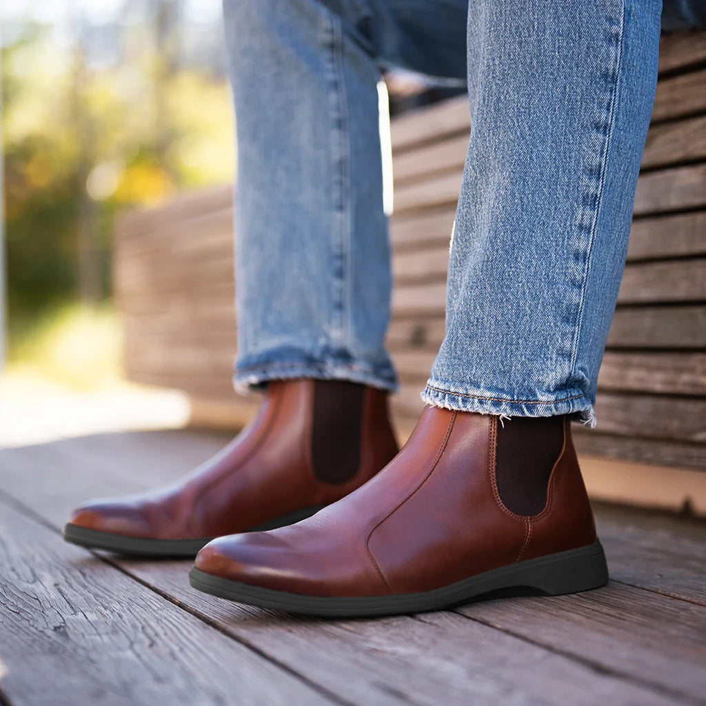 Brown leather boots worn with blue jeans on a wooden floor.