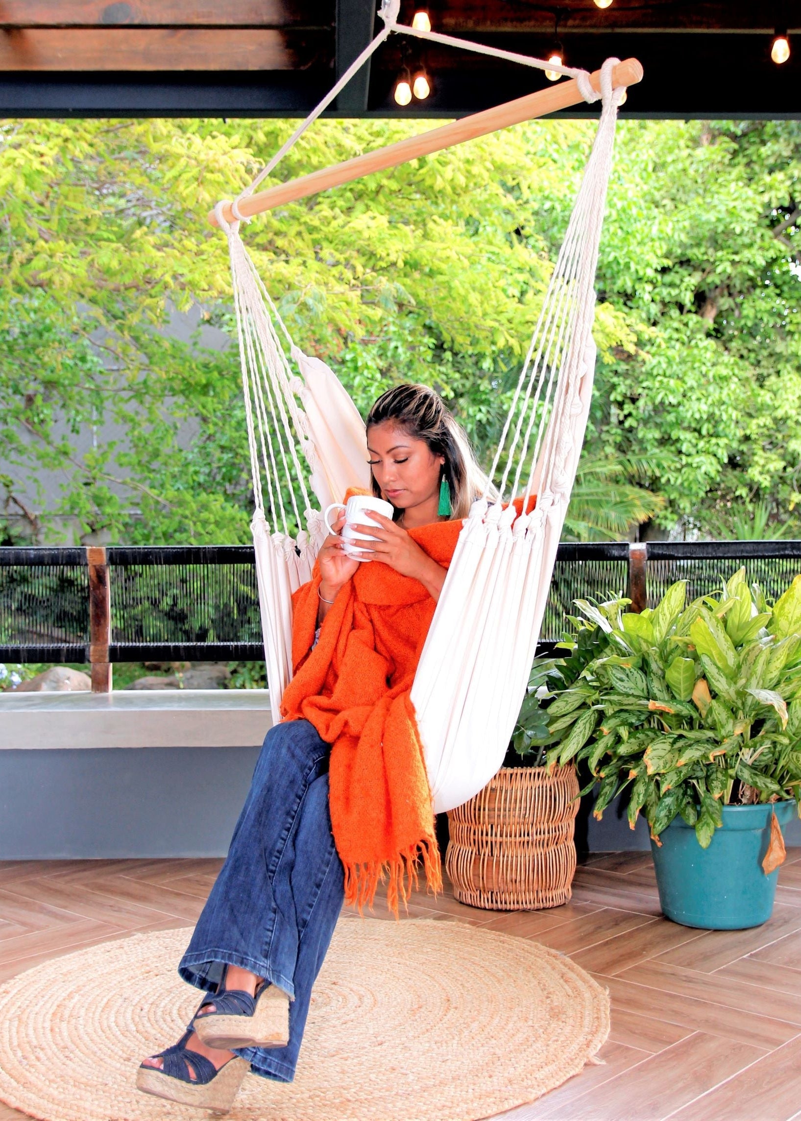 Person sitting in a white hammock on a wooden deck with plants and a railing in the background