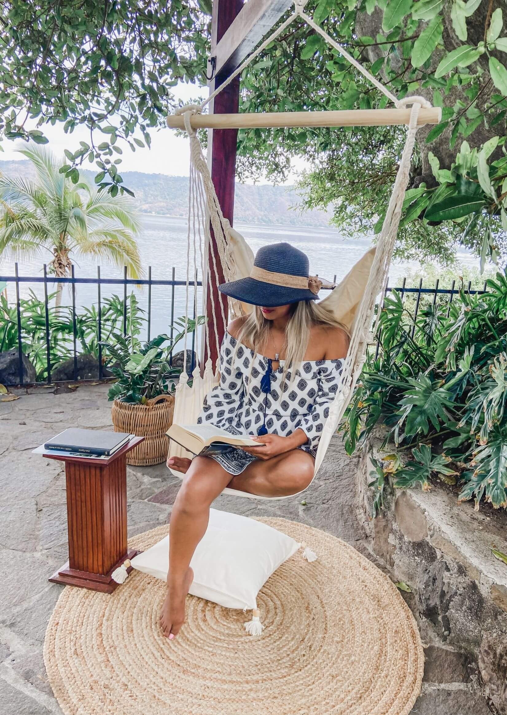 Woman reading a book in a hammock by a waterfront with greenery.