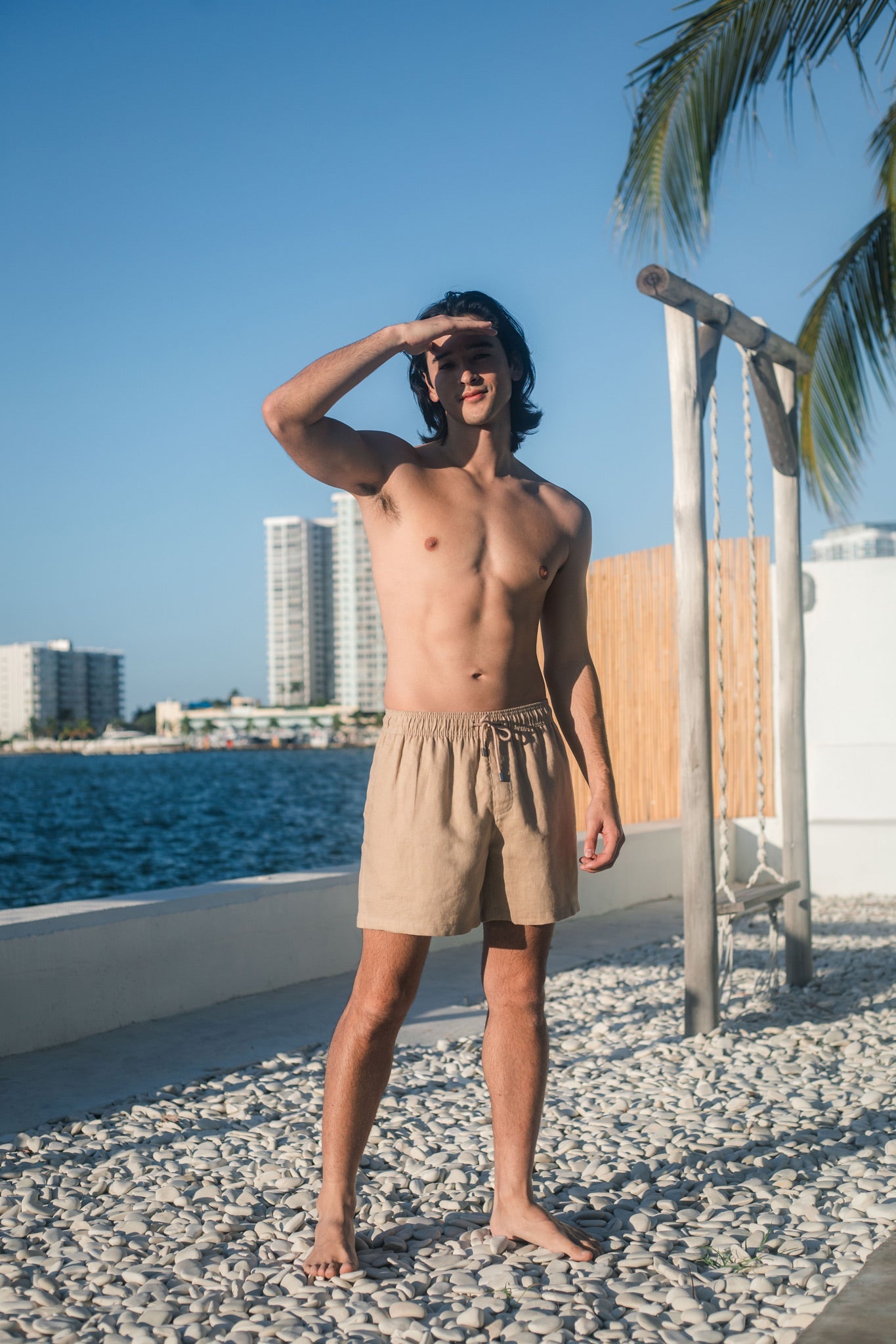 Man in beige shorts standing by a waterfront with palm trees and buildings in the background.