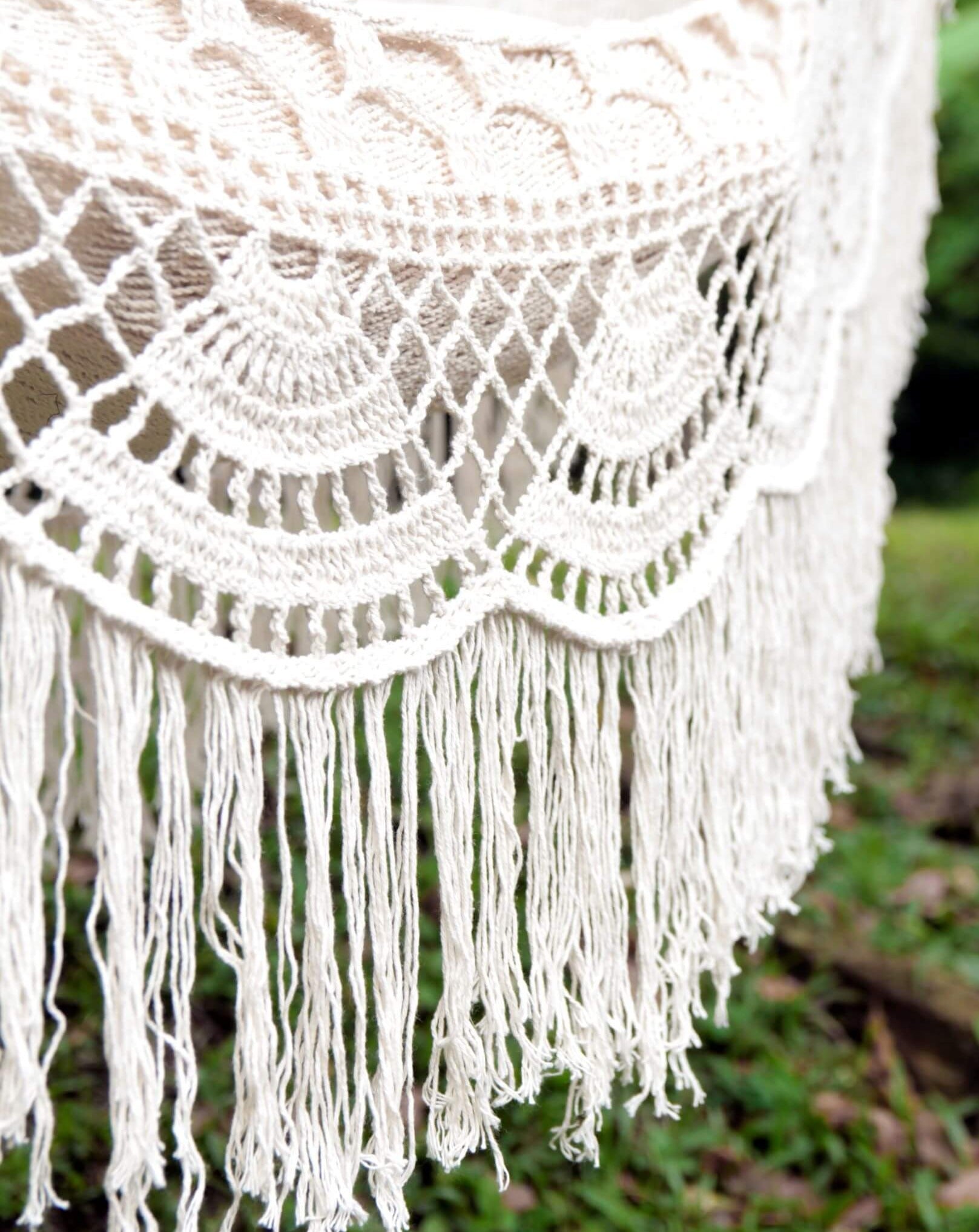 Close-up of a white crochet blanket with fringe against a blurred natural background