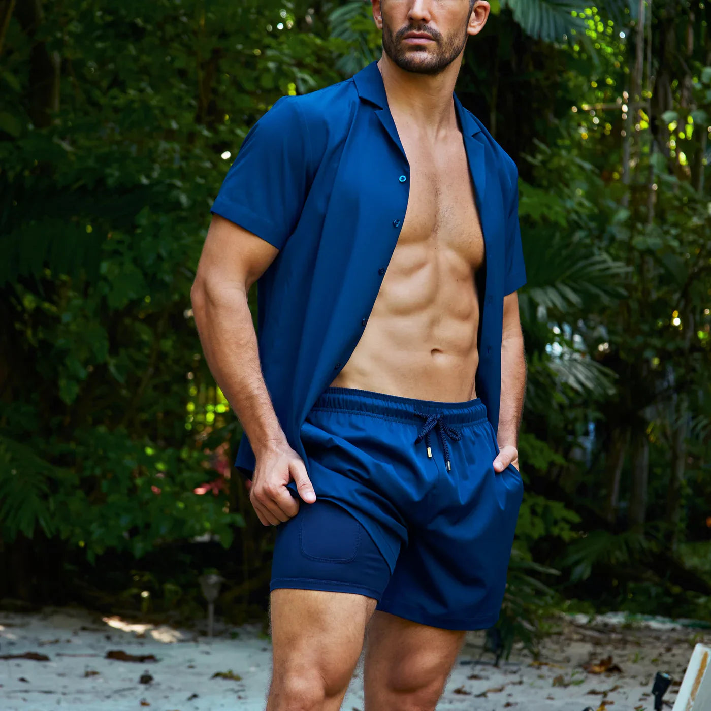 Man wearing a blue shirt and shorts standing outdoors with greenery in the background
