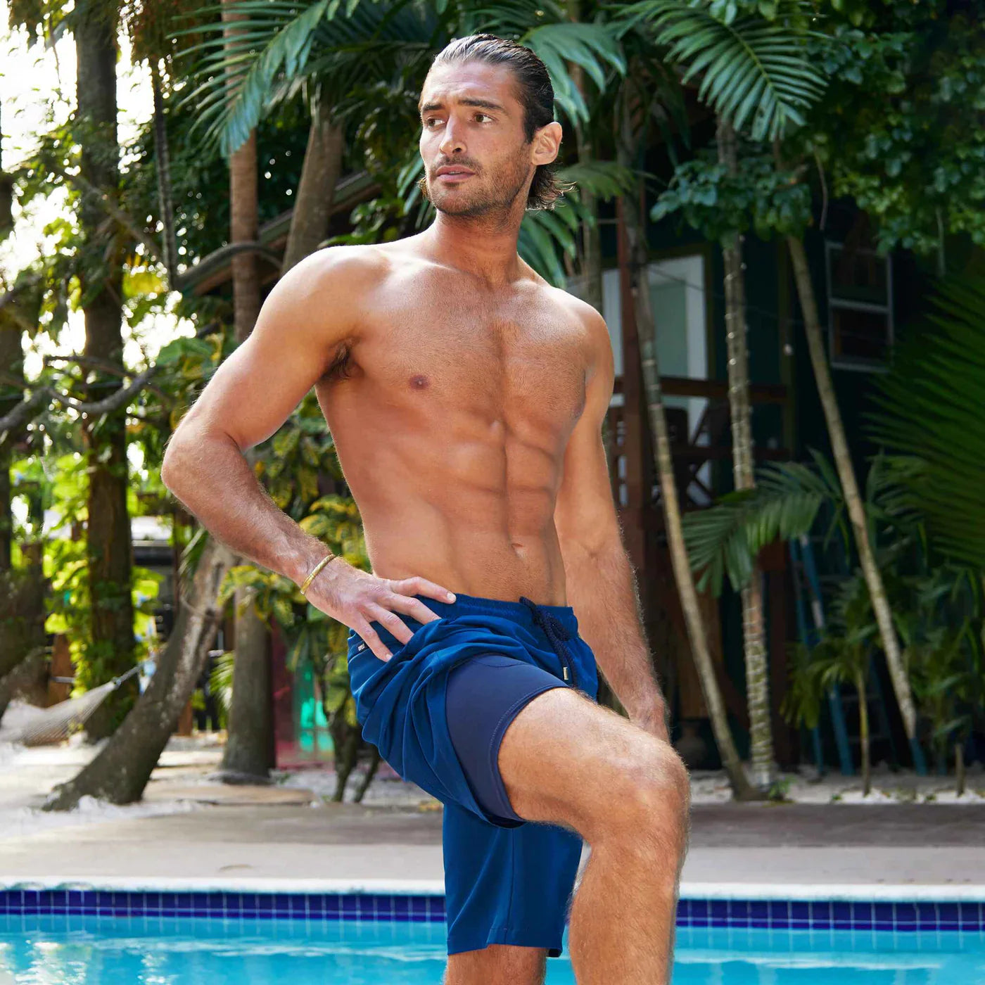 Man in blue swim shorts standing by a pool with greenery in the background