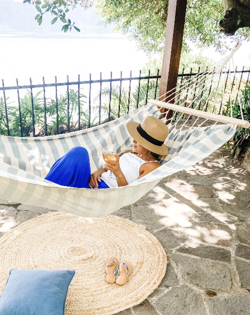 Person relaxing in a striped hammock with a drink, surrounded by greenery.
