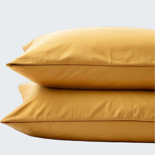Two mustard yellow pillows stacked on a bed with a wooden headboard and blurred window in the background.