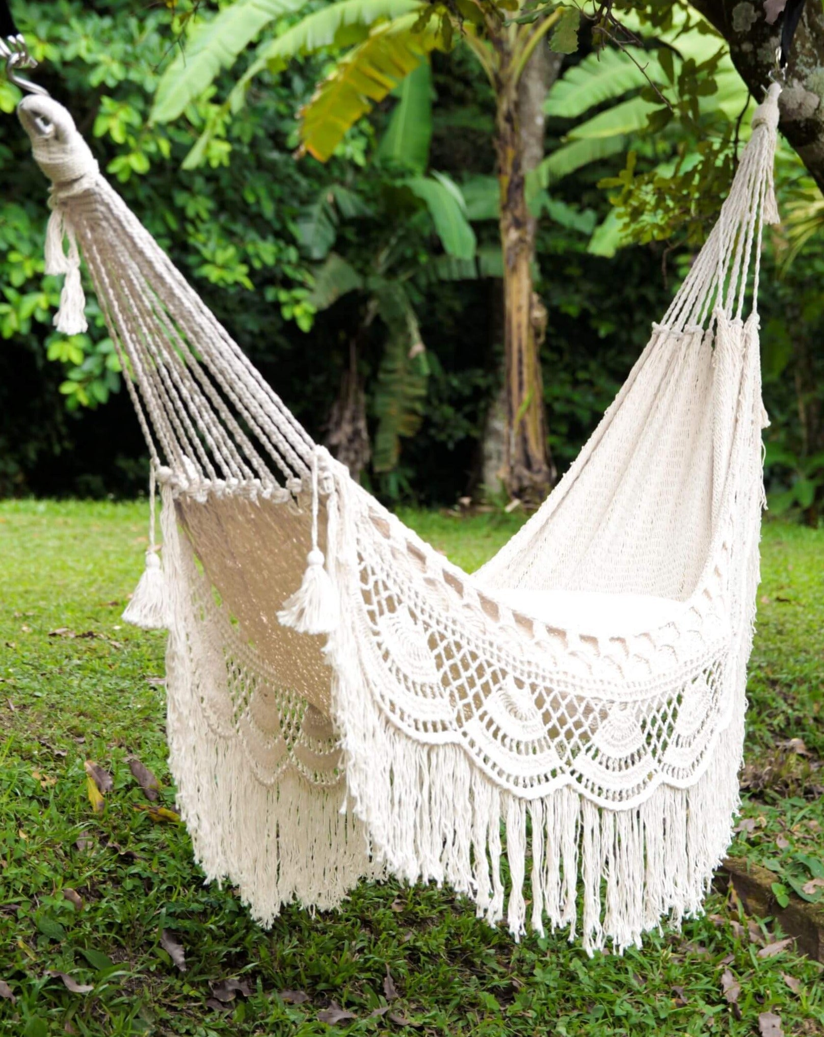 White hammock with tassels hanging in a natural setting with greenery.