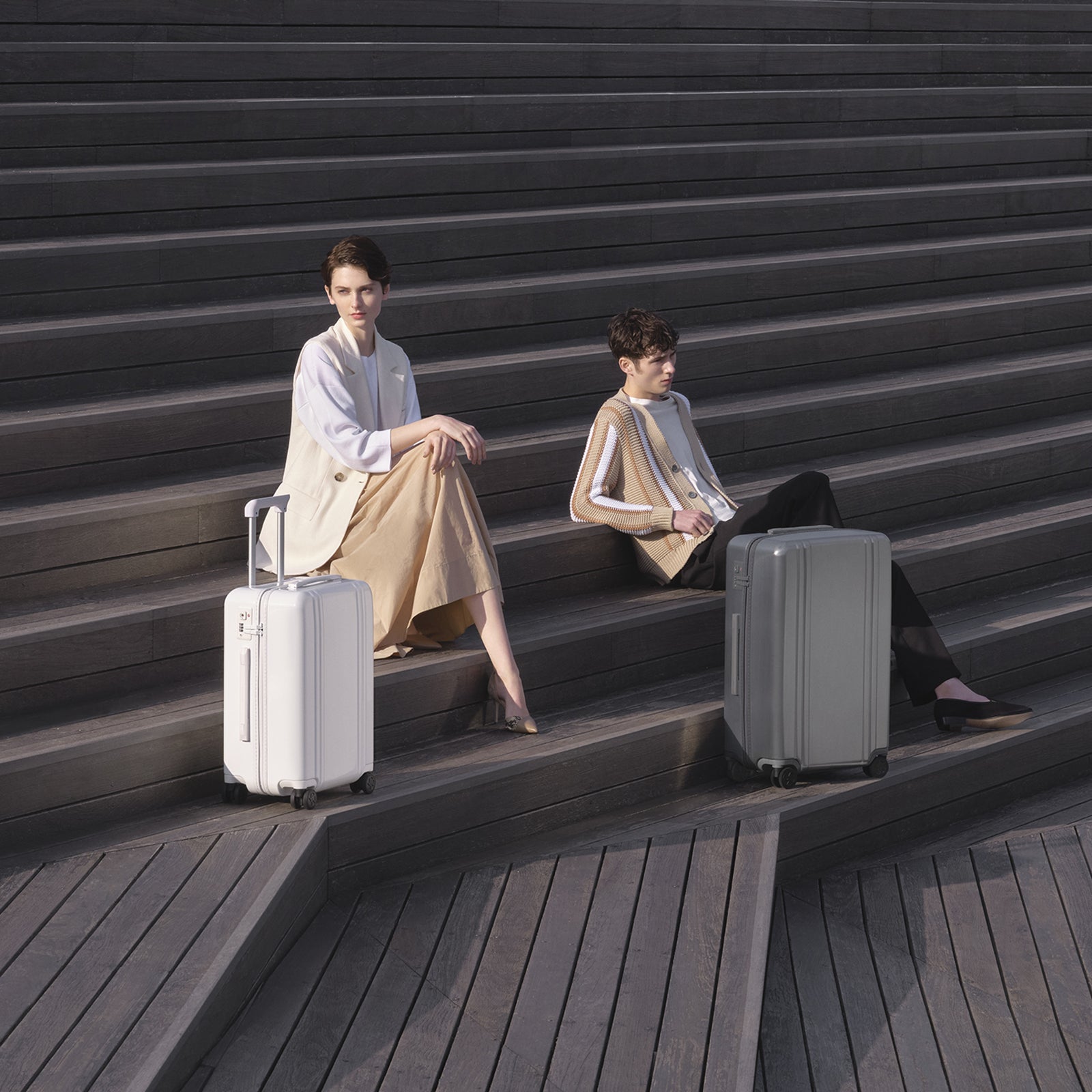 Two people sitting on steps with suitcases, one with a white suitcase and the other with a gray suitcase.