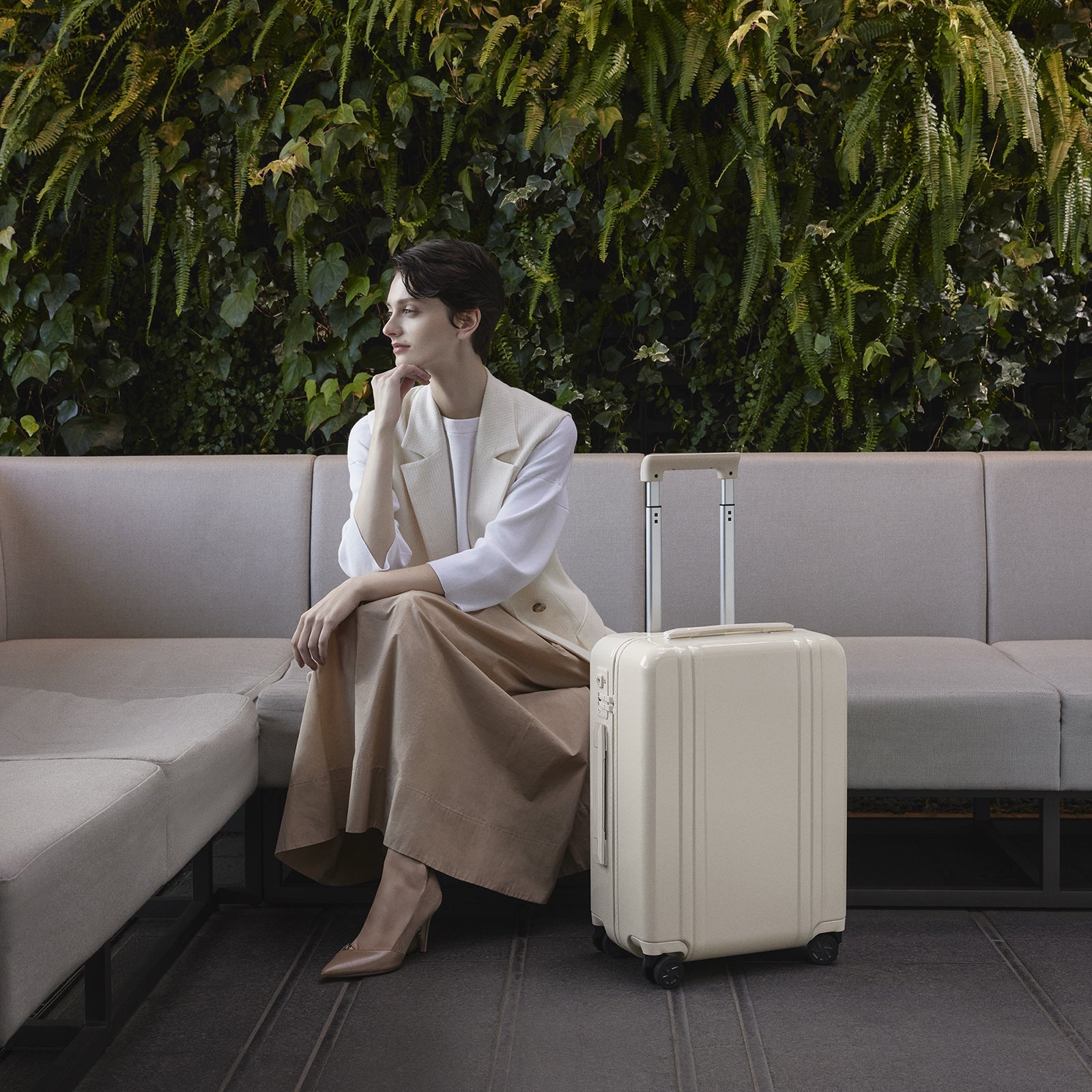 Woman sitting on a couch with a beige suitcase next to her against a green foliage background
