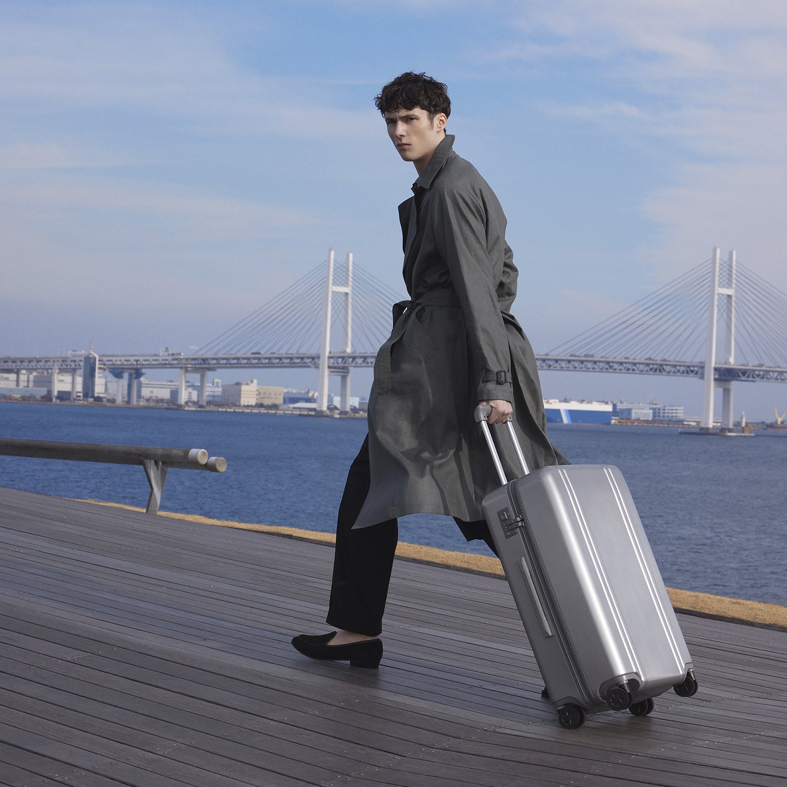 Man in a gray coat pulling a silver suitcase on a wooden deck with a bridge and water in the background